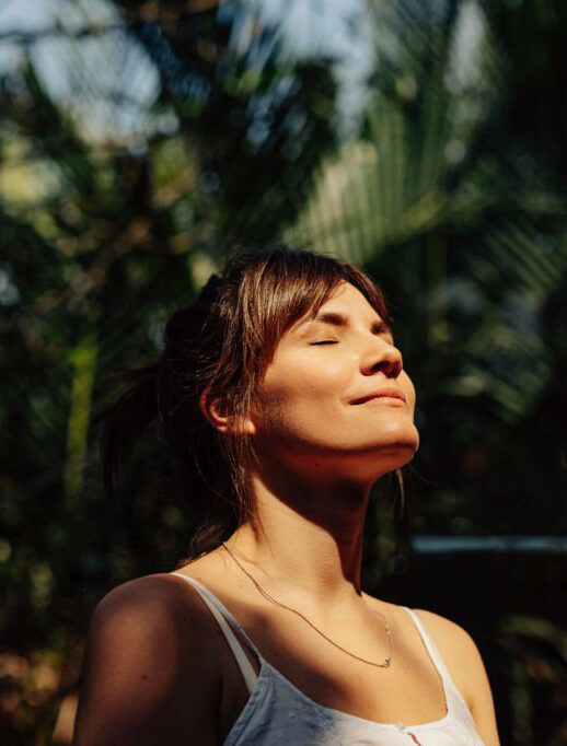Close up upper body shot of a beautiful happy young Caucasian woman enjoying the warm sunlight and tropical atmosphere with her eyes closed surrounded by palm trees in a tropical public park.